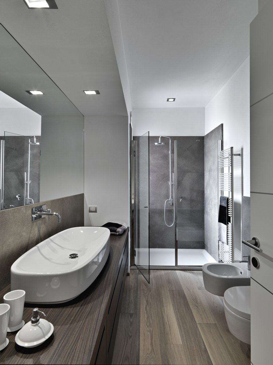 Bagno moderno lungo e stretto con water e bidet. Lo specchio aumenta la larghezza della stanza, così come la chiusura in vetro della doccia. Pavimenti e mobile in legno.
