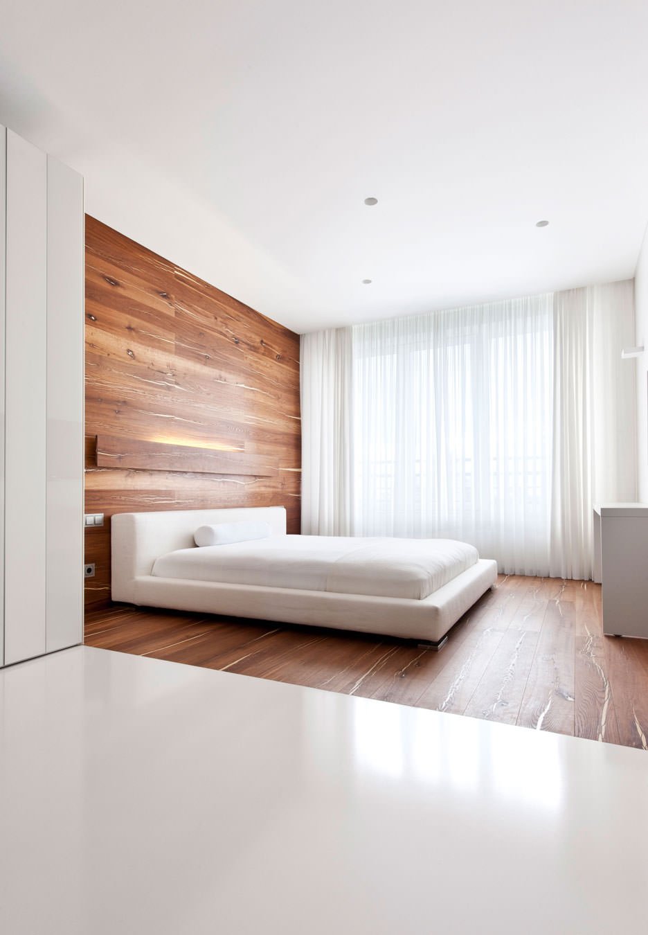 Il pavimento si prolunga su una porzione di parete differenziando la zona letto dal resto dell’ambiente. Stile moderno minimal - colore bianco in contrasto con il legno