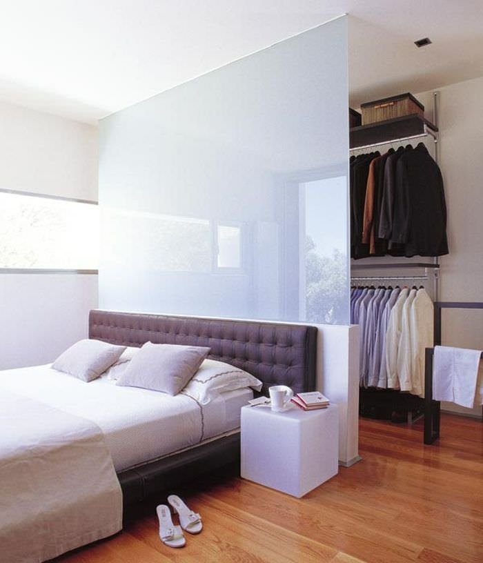 Stupenda camera da letto moderna in cui la testiera in vetro temperato crea una cabina armadio. Il mobilio fa l’architettura della stanza. Pavimento in legno.