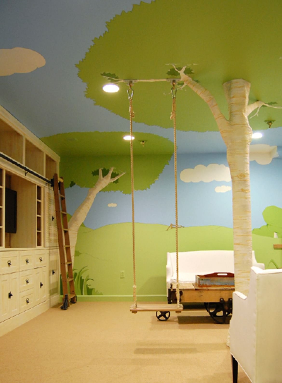 Originale e moderna cameretta per bambini, con altalena,  dipinta con elementi naturali: prato, alberi e cielo. Un vero albero è stato posizionato nella stanza per dare un aspetto più veritiero al contesto