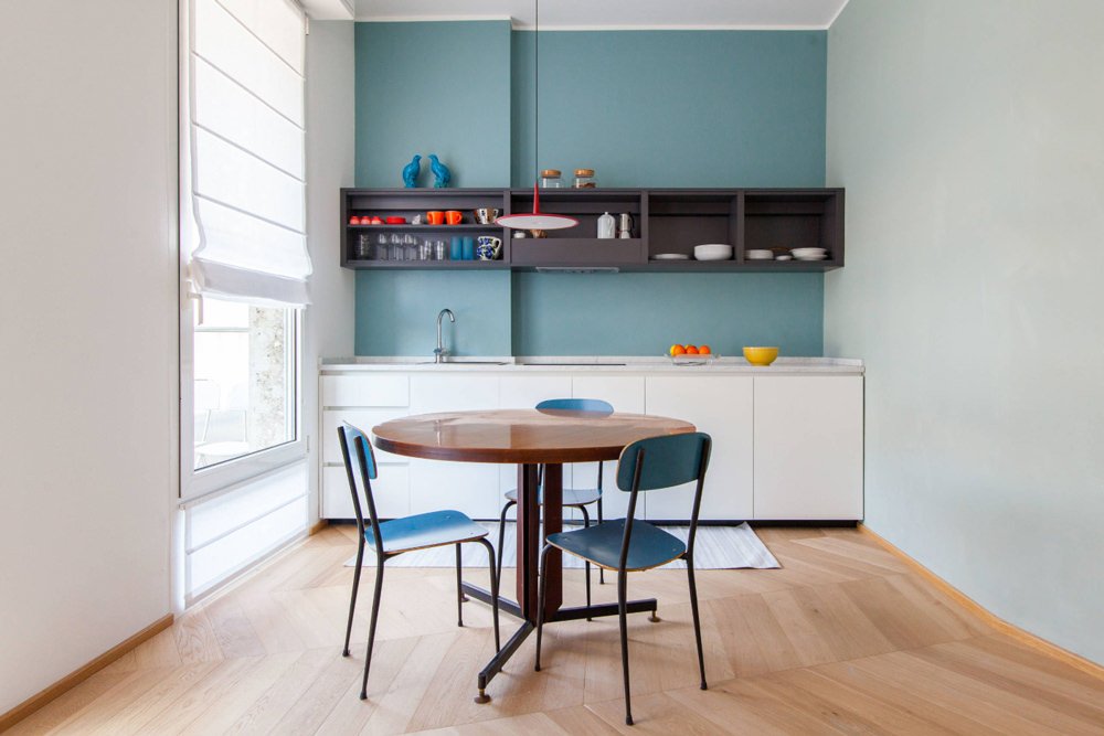 Cucina piccola moderna e funzionale con le pareti, le sedie ed alcuni oggetti decorativi di colore blu. Pensile marrone scuro, mobile bianco ed alcune decorazioni di colore rosso per dare contrasto e vivacità allo spazio. Pavimenti in legno.