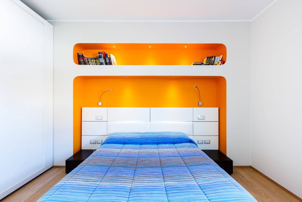 Moderna camera da letto con muro retrostante in cartongesso in cui sono state create delle nicchie, colorate in arancione, per testiera e libreria.