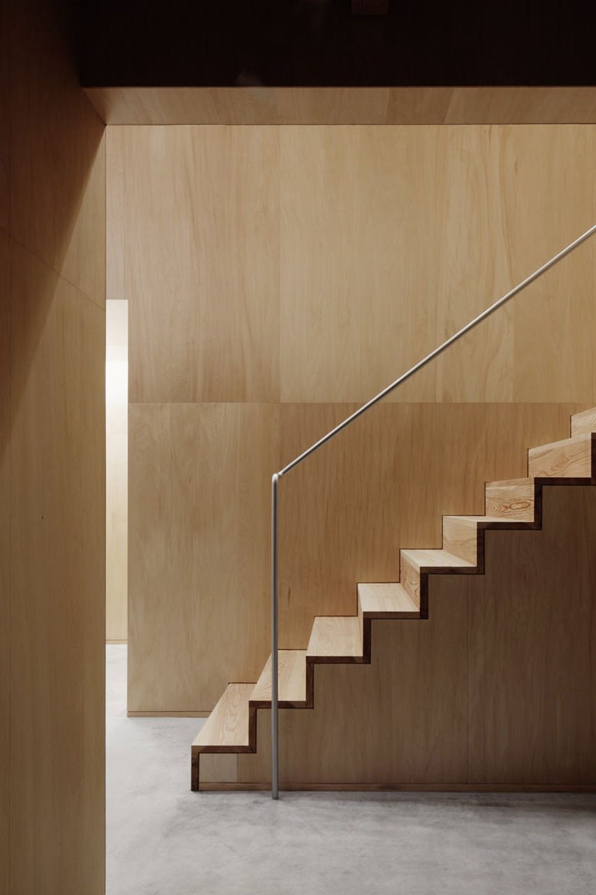 Scala in legno minimalista mimetizzata in una stanza completamente rivestita in pannelli di legno. Spiccano solo i gradini e la semplice ringhiera metallica