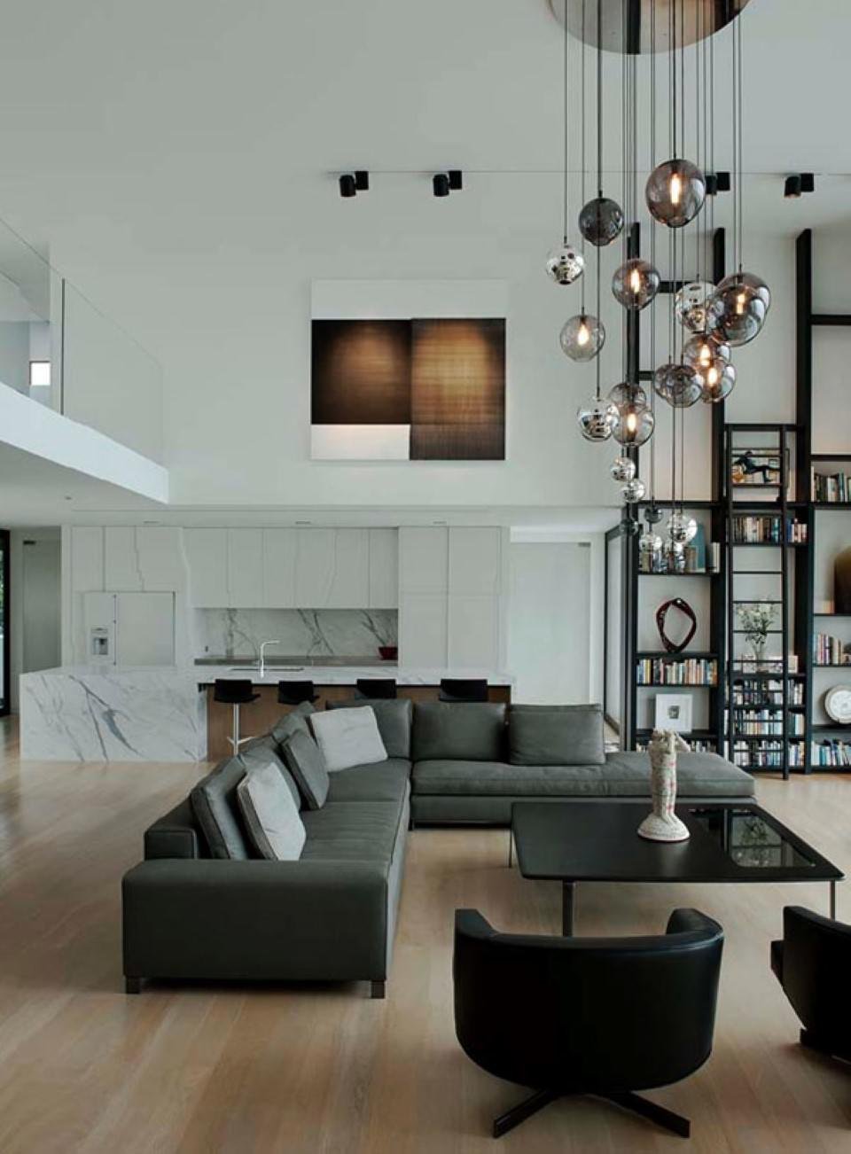 Soggiorno ultramoderno in un ambiente open space con soffitti molto alti. Arredamento, libreria e divani neri, pavimenti in legno chiaro e particolare illuminazione